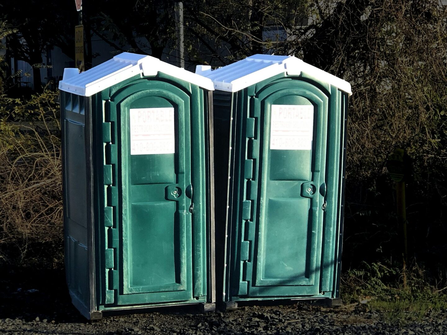 Two green portable toilets side by side in bright sunlight.