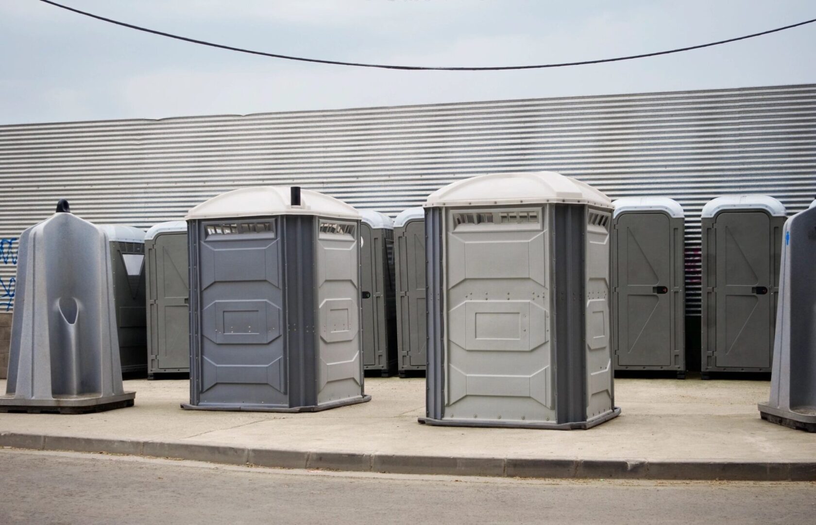 A row of portable toilets and a hand washing station in a parking area with a curved metal roof in the background.